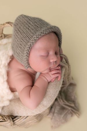 Peaceful sleeping newborn baby wearing a gray knit bonnet hat, nestled in a wicker basket with soft blankets.