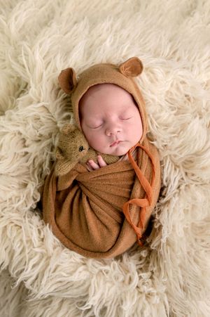 Sleeping newborn swaddled in brown wrap wearing bear bonnet, holding knit teddy bear on fluffy cream rug.