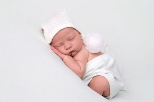Sleeping newborn baby wearing a white knit hat with pompom, wrapped in white, on a white background.