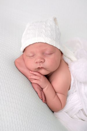 Sleeping newborn baby wearing a white knit hat, wrapped in white, resting peacefully on a white background.