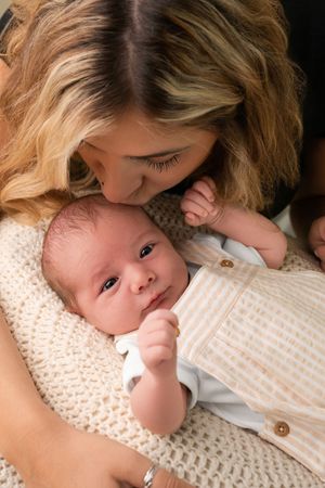 A mother tenderly kisses her newborn baby wrapped in a cream knit blanket wearing a striped onesie.