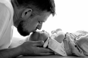 Black and white photo of a bearded father gently kissing his newborn baby lying on a white surface.