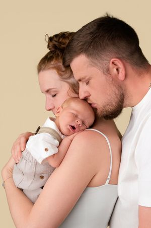 A young couple tenderly kisses their sleeping newborn baby, sharing a loving family moment against a soft beige background.