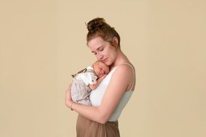 A mother tenderly holds her sleeping newborn baby close to her chest against a soft beige background.