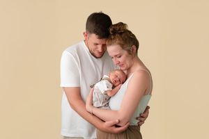 Young parents tenderly cradling their sleeping newborn baby against a soft beige studio background.