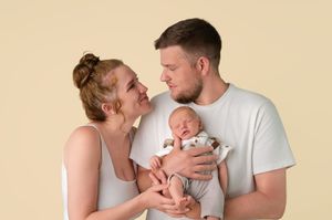 Young couple in white tops gazing at each other while holding their sleeping newborn baby against beige background.