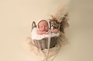 Newborn baby sleeping peacefully in a wicker basket with a teddy bear and fluffy white blanket.