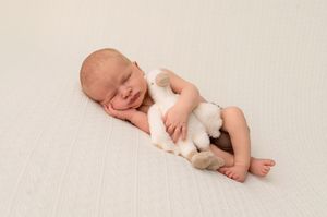 Newborn baby sleeping peacefully on white blanket, cuddling a small white stuffed animal toy.
