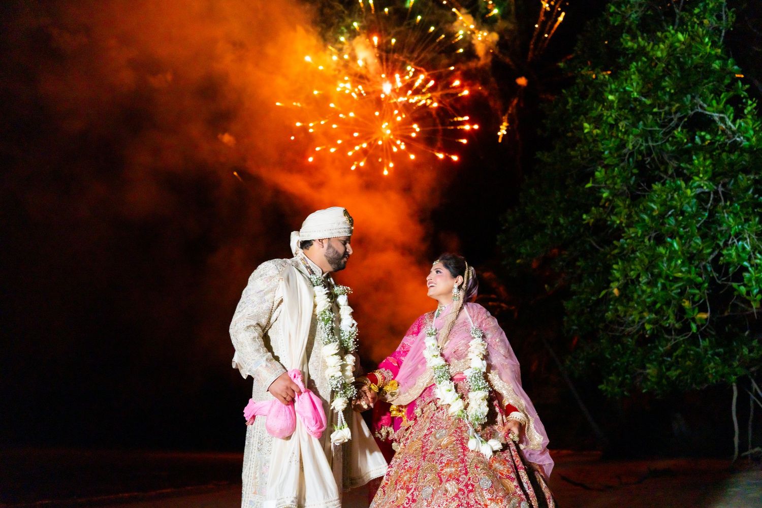 Indian couple in traditional wedding attire celebrate with fireworks display behind them at night.