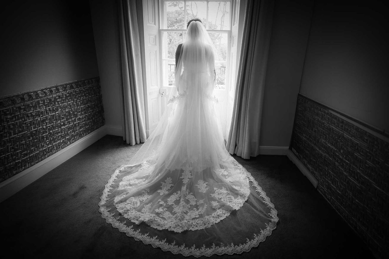 Black and white photo of a bride in a lace wedding gown and veil standing before a sunlit window.
