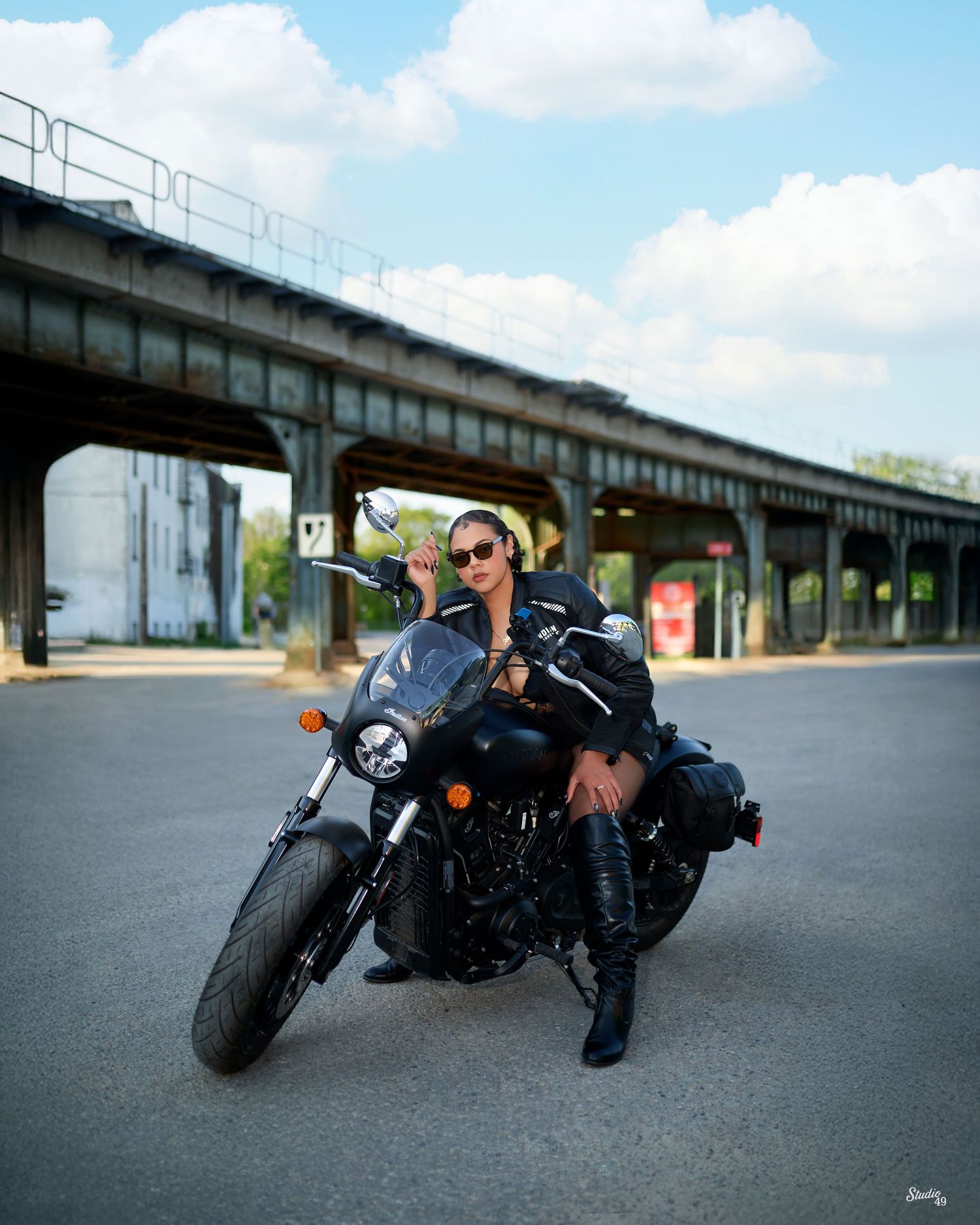 A woman in leather jacket and boots poses confidently on a black motorcycle under an urban overpass on a sunny day.