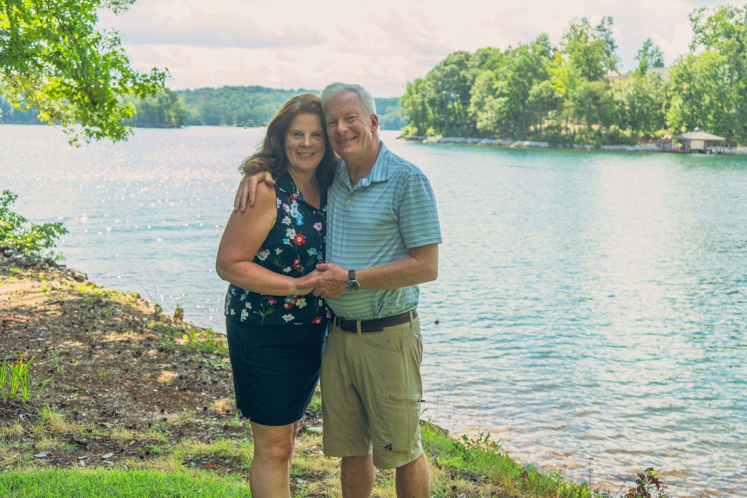 Couple smiling and embracing by a serene lake surrounded by lush green trees on a cloudy day.