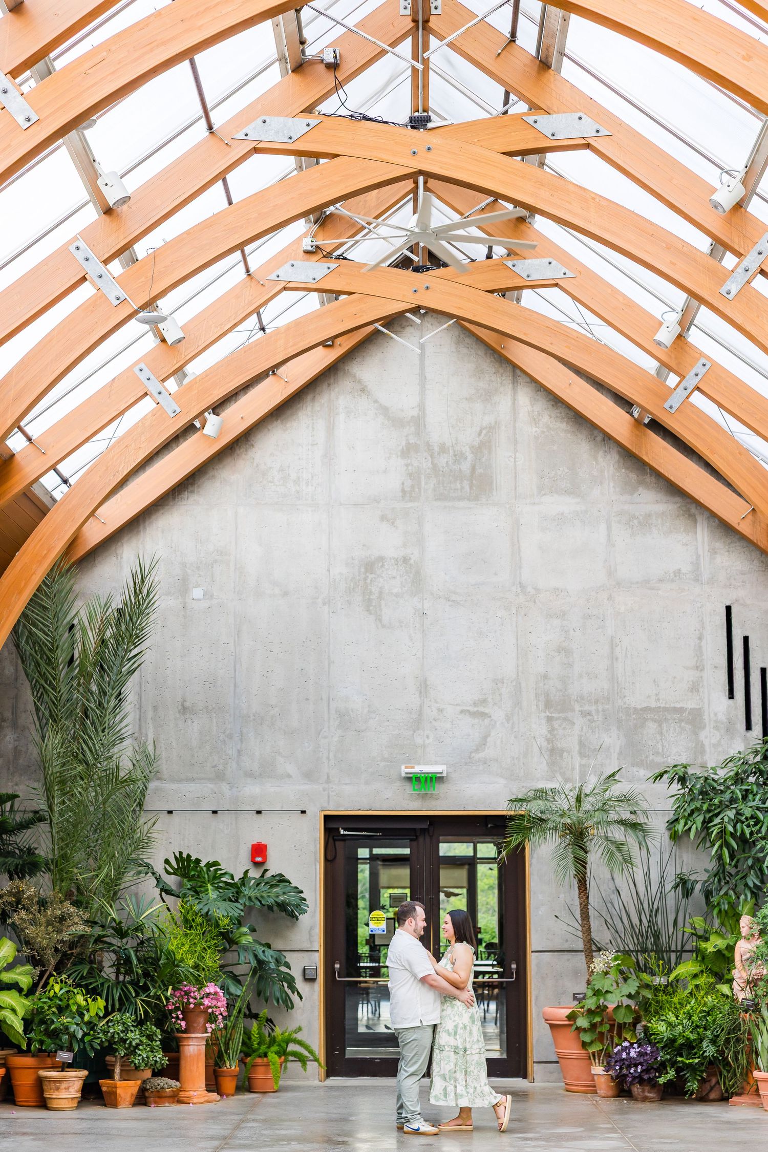 Couple embraces under stunning arched wooden ceiling in a greenhouse venue surrounded by lush tropical plants.