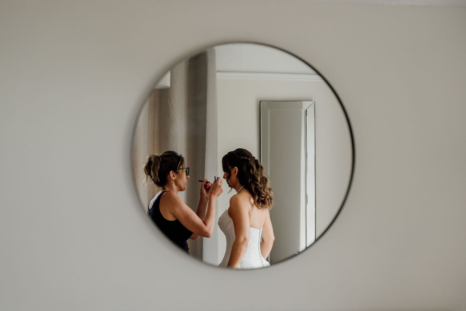A makeup artist applies makeup to a bride in a white wedding dress, reflected in a round mirror on a white wall.