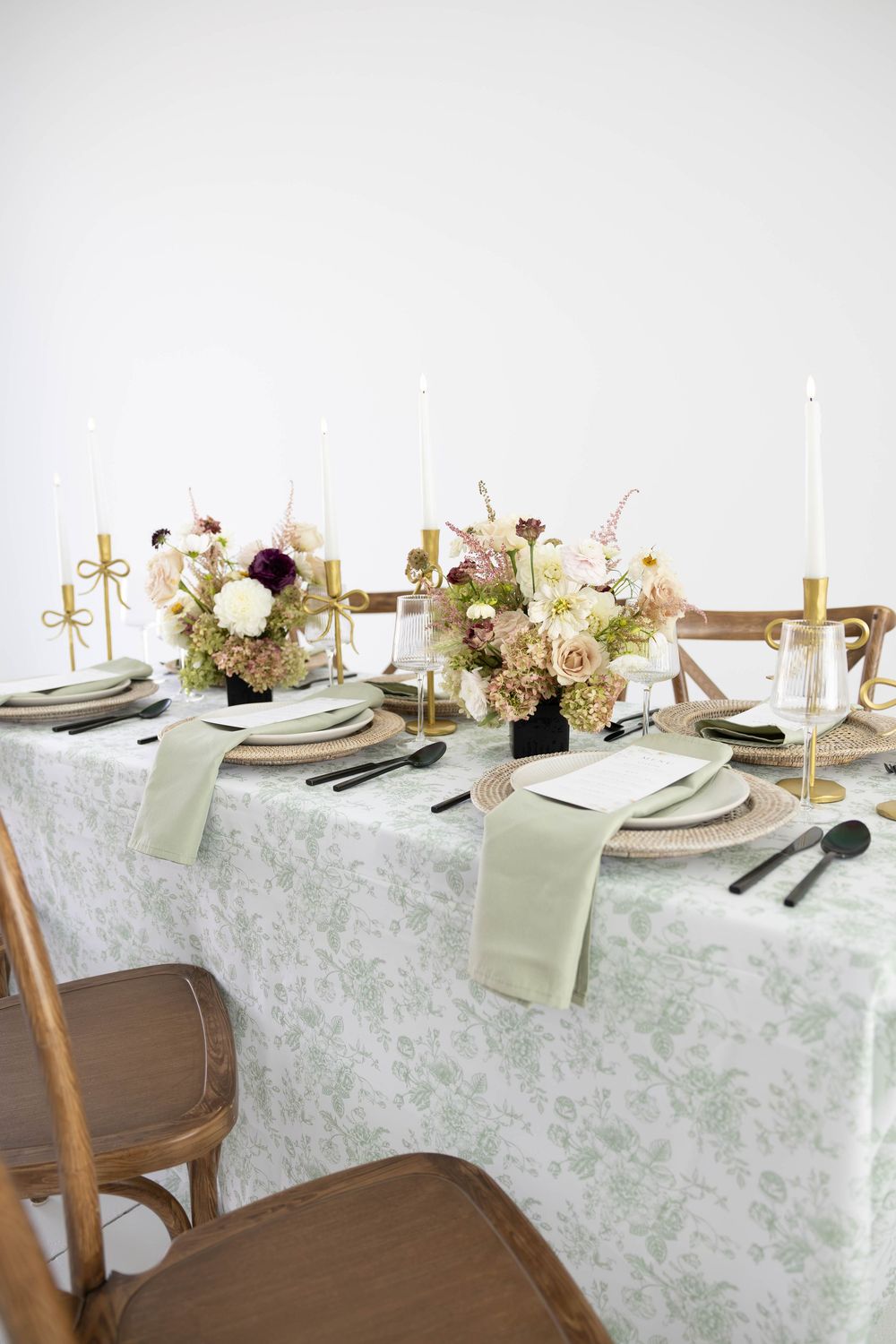 Elegant wedding tablescape with sage green floral tablecloth, gold candlesticks, woven chargers, and lush floral centerpieces.