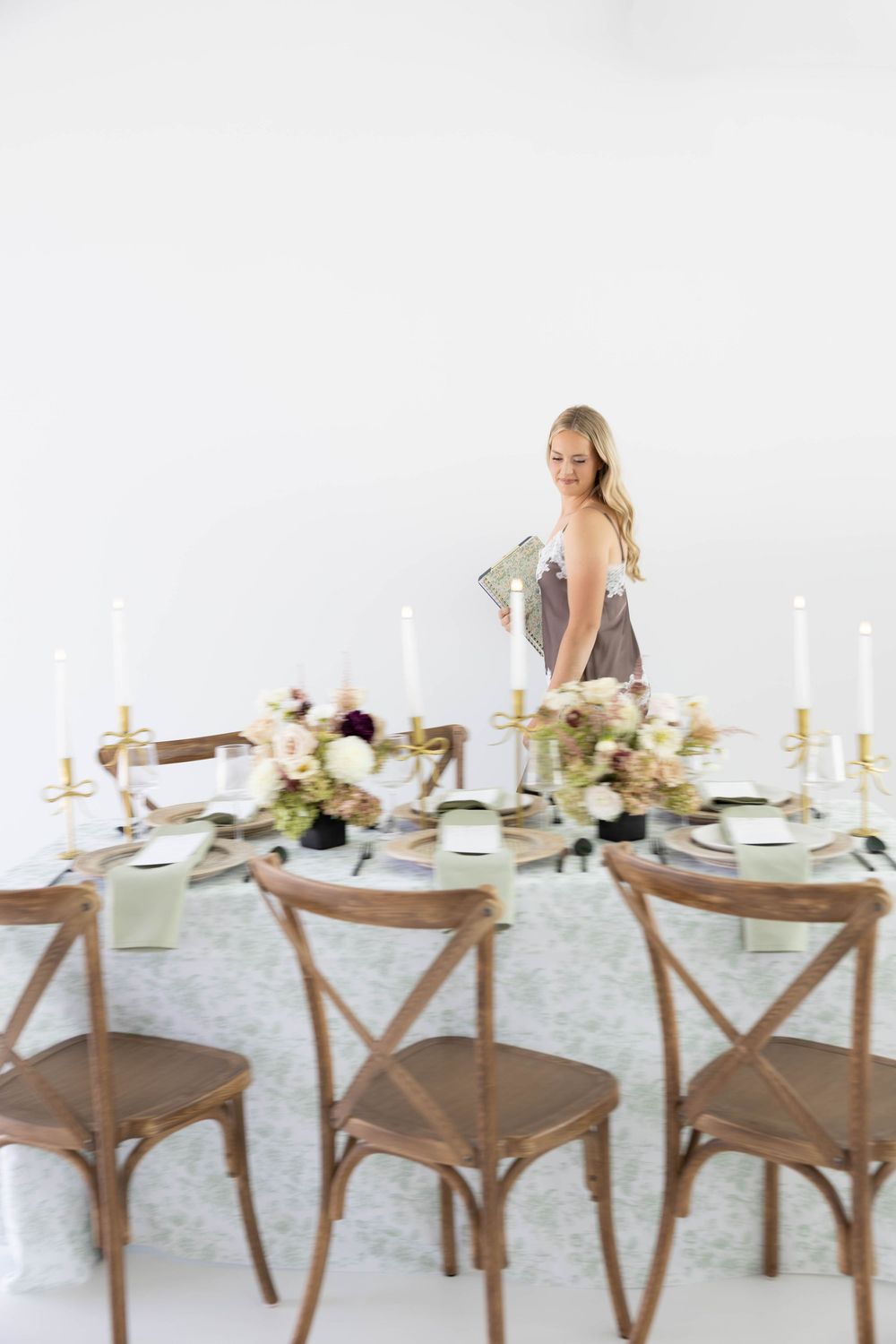 Elegant wedding table setting with floral centerpieces, taper candles, and wooden cross-back chairs, with a blonde woman in background.
