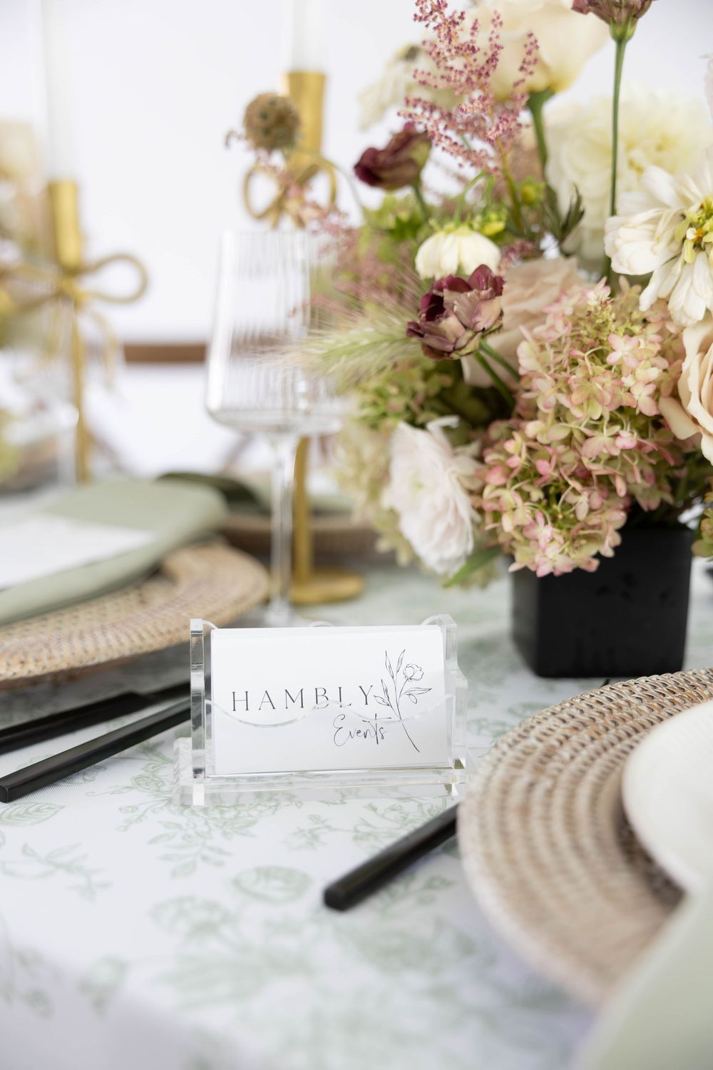 Elegant wedding table setting with floral centerpiece, gold candles, woven placemats, and Hambly Events place card.