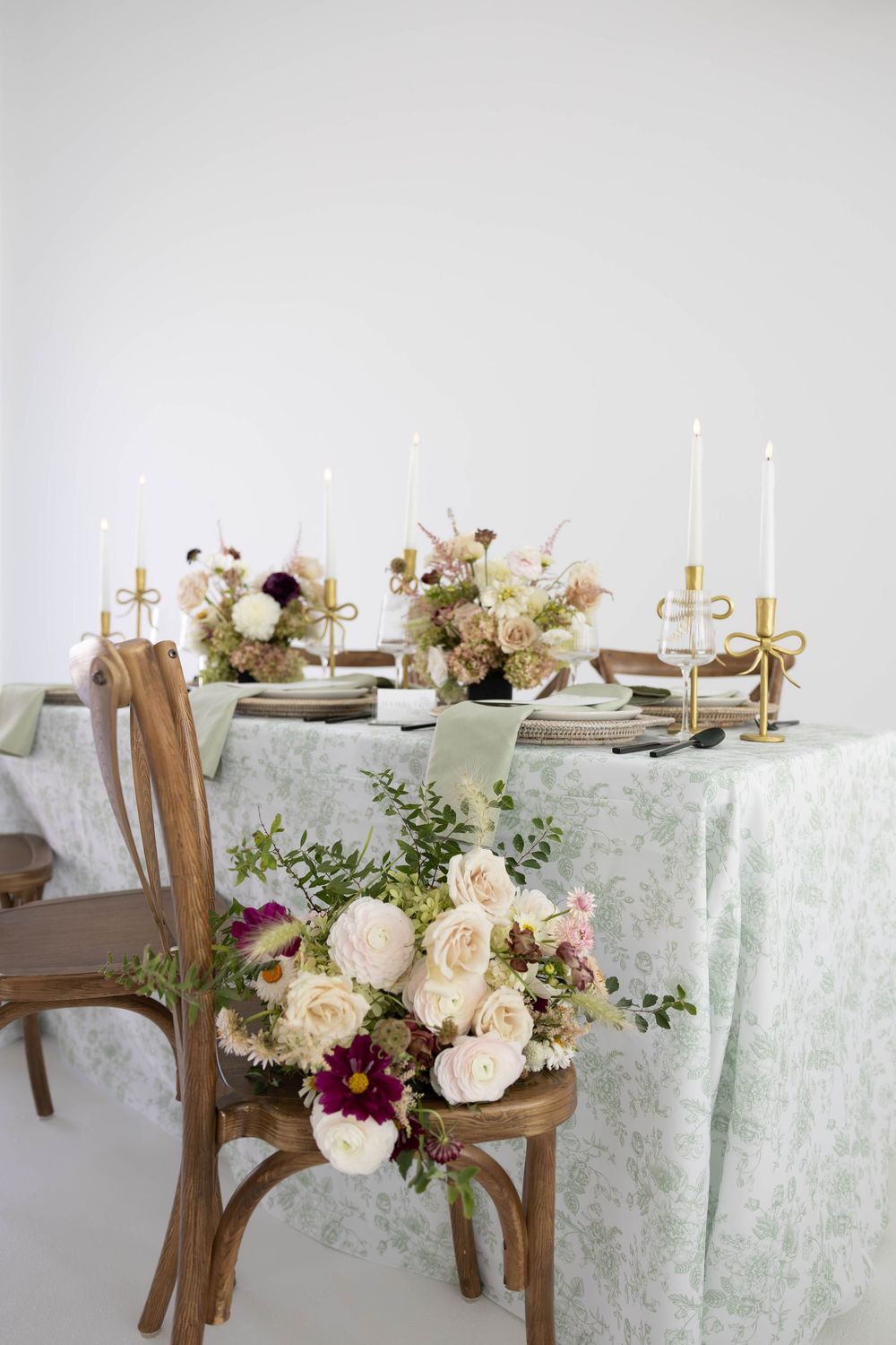 Elegant wedding tablescape with mint green floral linen, gold candlesticks, and cream rose bouquet on wooden chair.