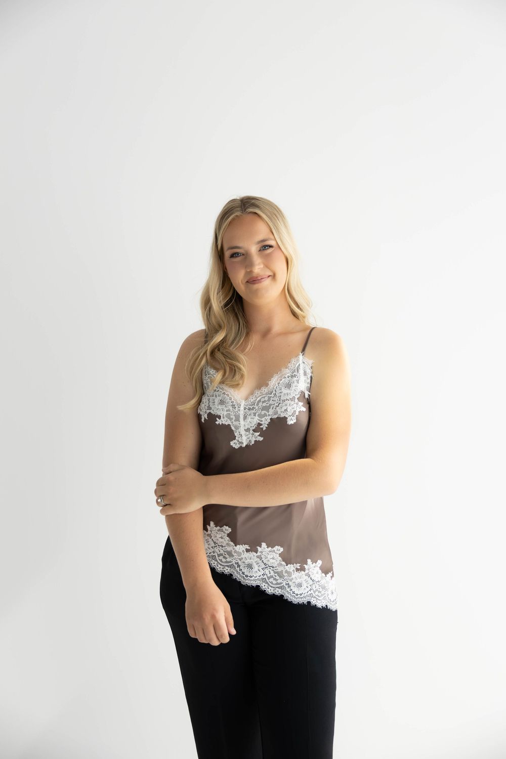 Woman wearing a brown satin camisole with white lace trim detailing, paired with black pants on white background.