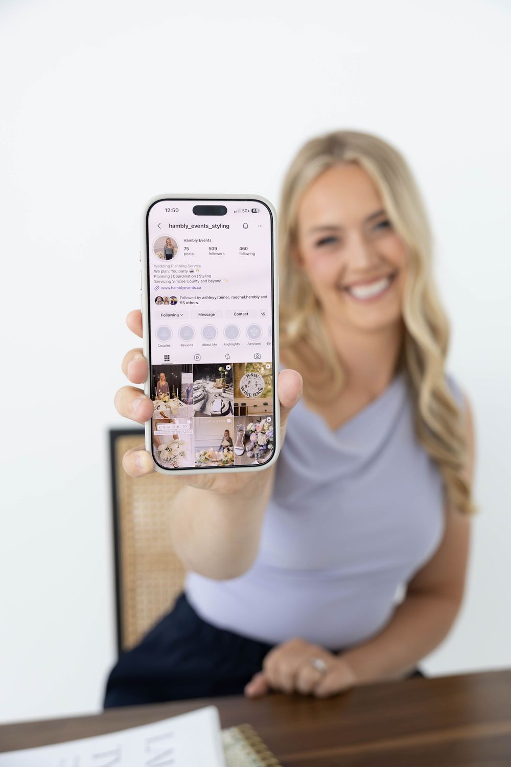 Smiling blonde woman holding smartphone showing Instagram profile page at desk.