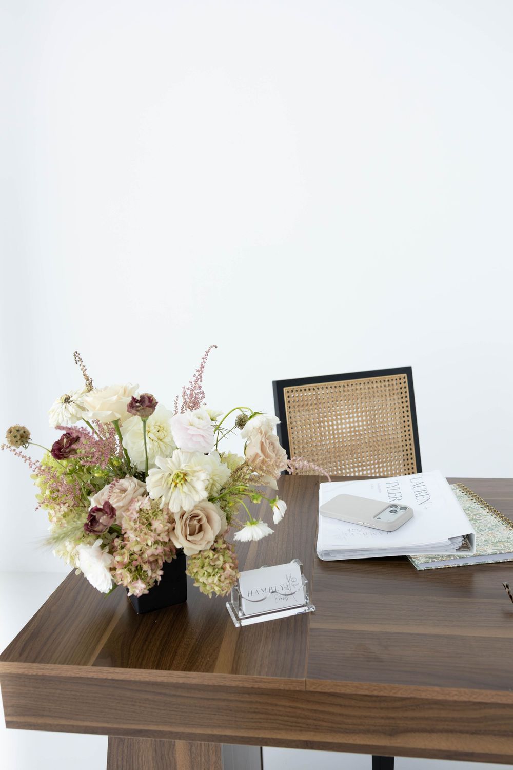Elegant wooden desk with floral arrangement, open notebook, and woven chair in a bright minimalist office setting.
