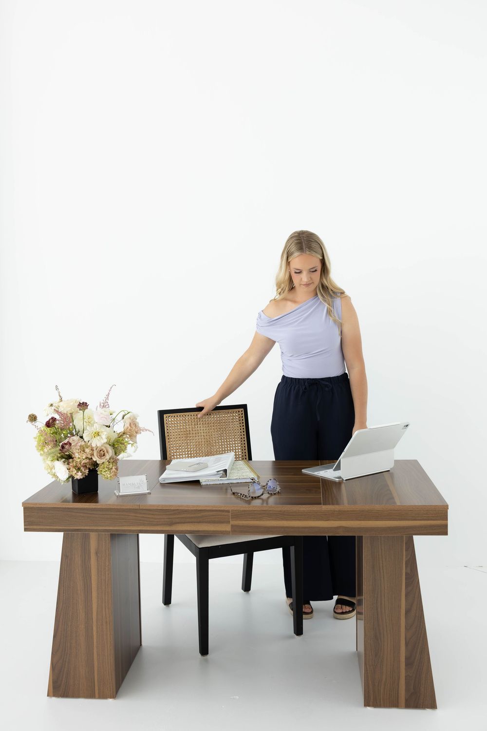 Blonde woman in blue top and navy pants standing at a modern walnut desk with laptop, chair, and floral arrangement.