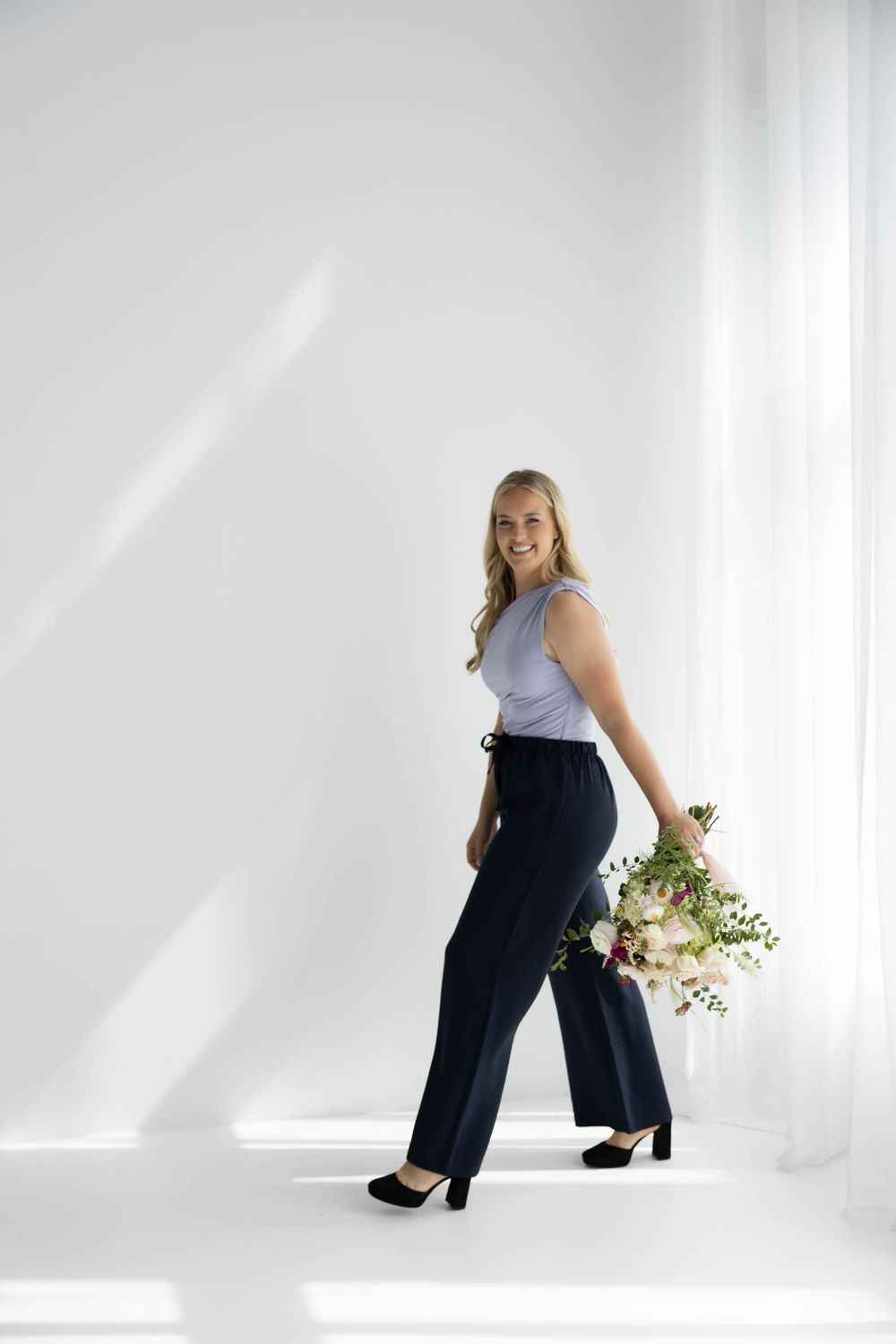 Smiling blonde woman in navy wide-leg pants and gray top holding a bouquet of pink flowers in a bright white studio.