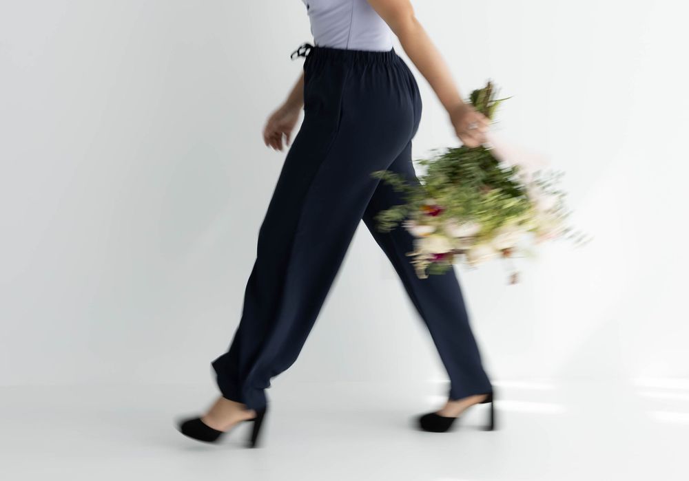 Woman in navy trousers and black heels walking while holding a bouquet of flowers against a white background.