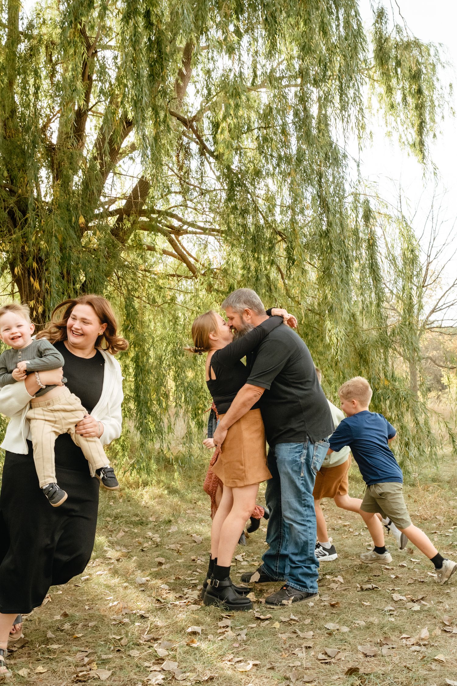 Family of four posing outdoors near willow trees in autumn, parents standing behind two young red-haired boys.