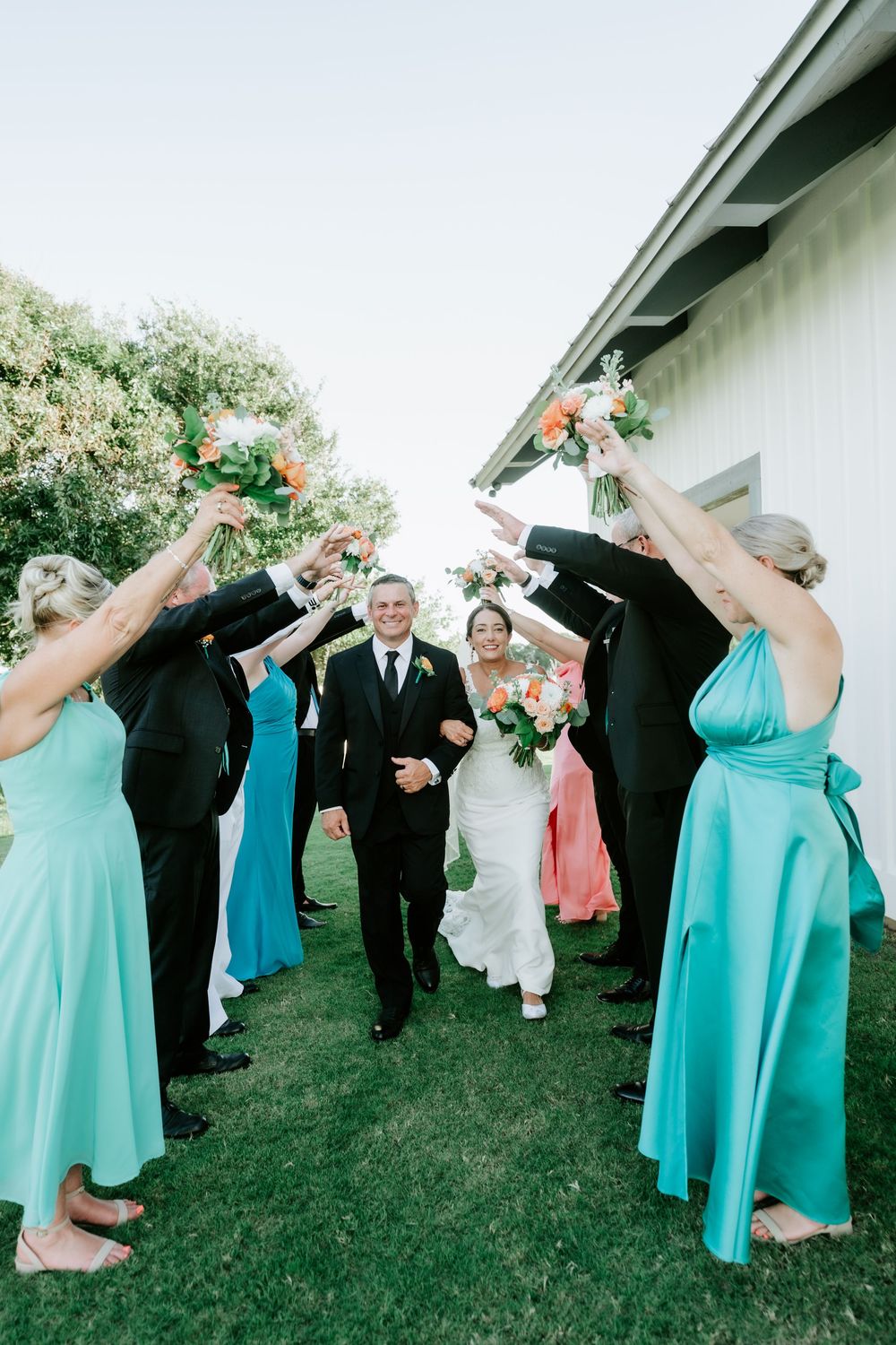 Bride and groom walk through an arch of raised bouquets held by wedding party in teal and black attire outdoors.