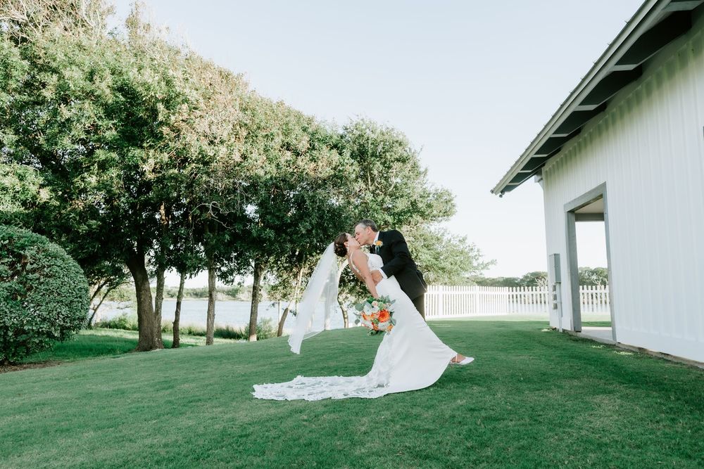 Bride and groom share a romantic dip kiss on lush green lawn outside white wedding venue surrounded by trees.
