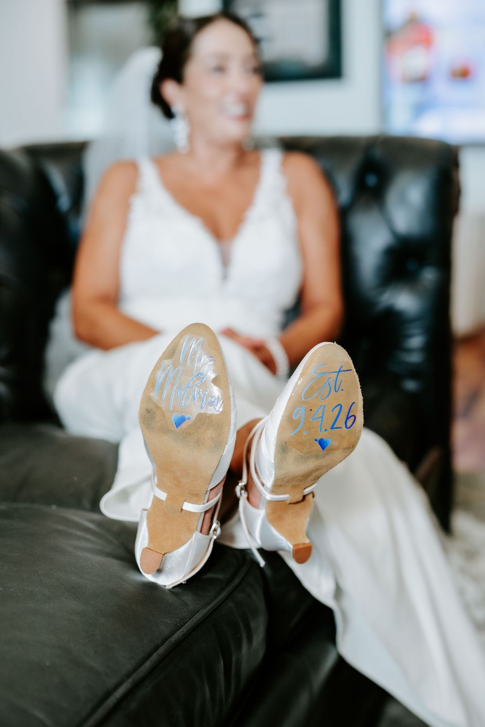 Bride in white wedding dress showing shoe soles with wedding date written on them while sitting on black leather sofa.