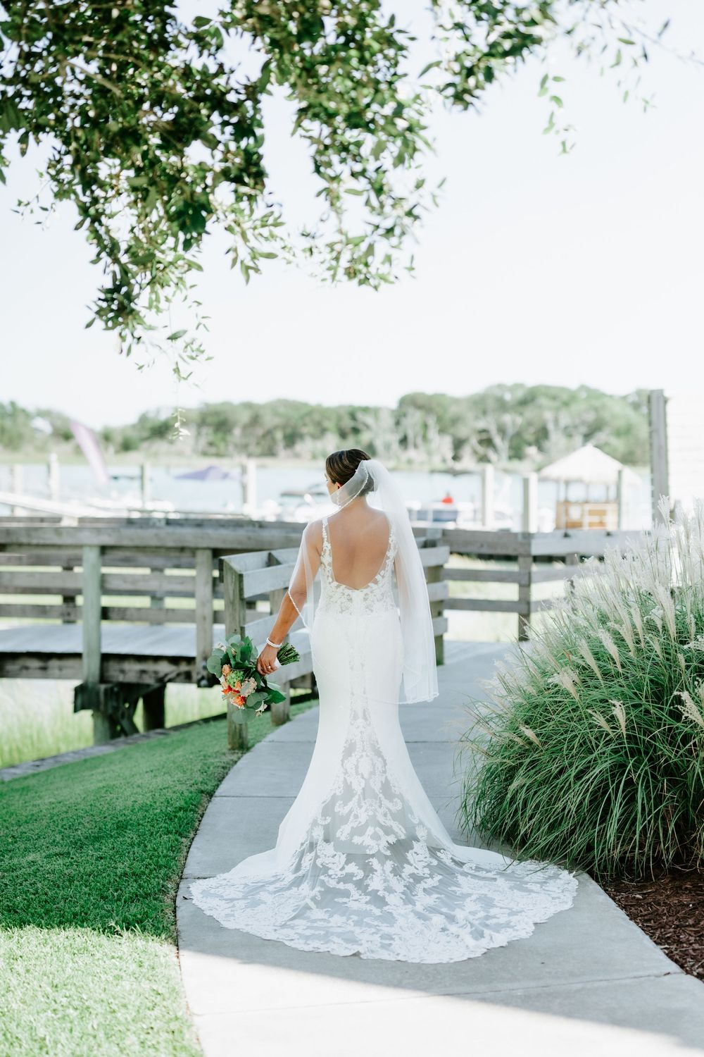Bride in lace mermaid gown with cathedral train walks along waterfront path holding bouquet, back detail visible.