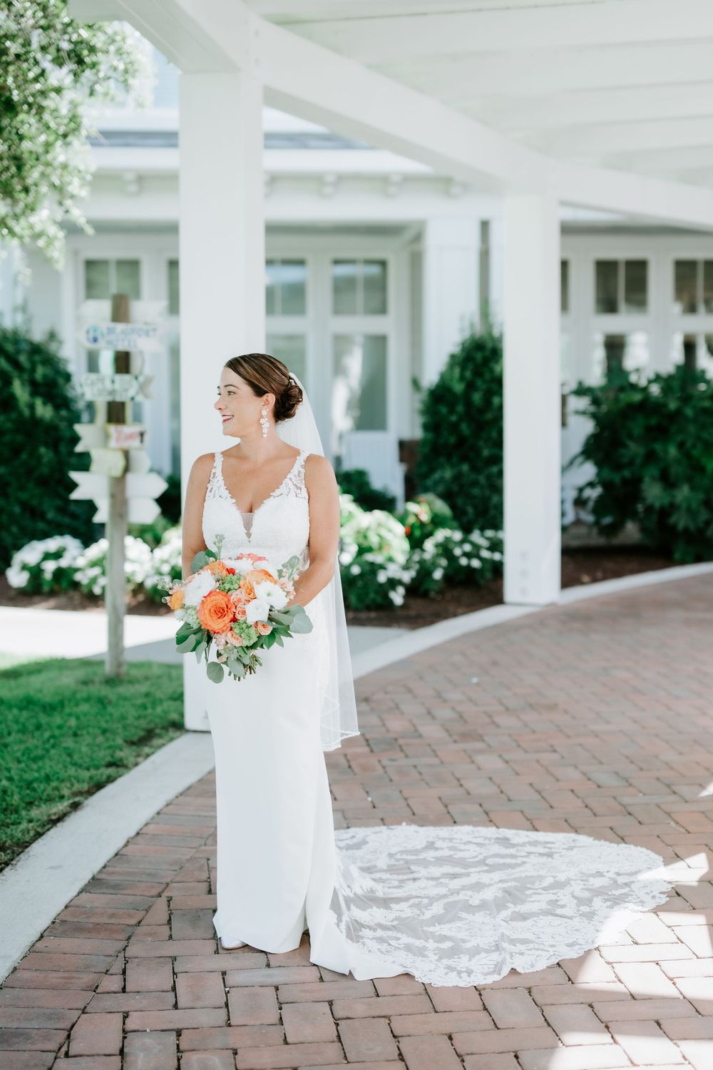 Smiling bride in white lace gown holding coral bouquet stands on brick path outside elegant white venue.