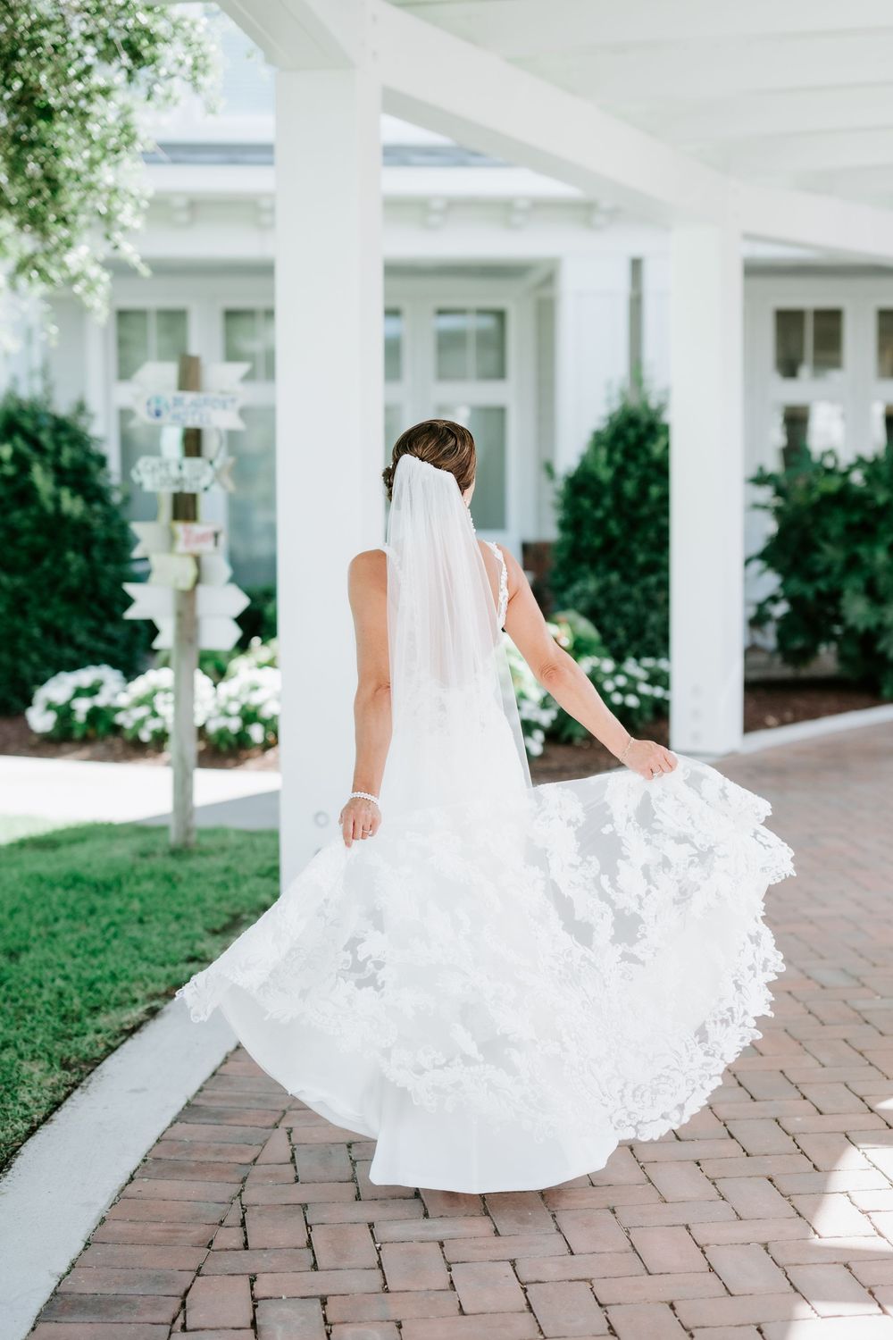 Bride in white lace cathedral veil and gown twirls on brick path outside white colonial venue.