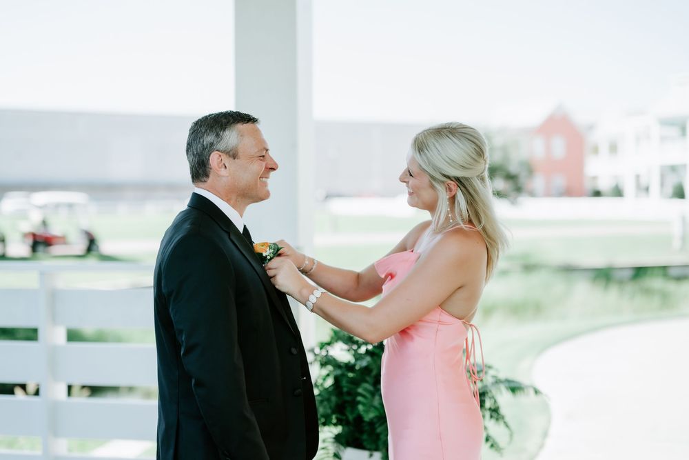 A woman in a pink dress pins a boutonniere on a man in a black tuxedo outdoors at a wedding.