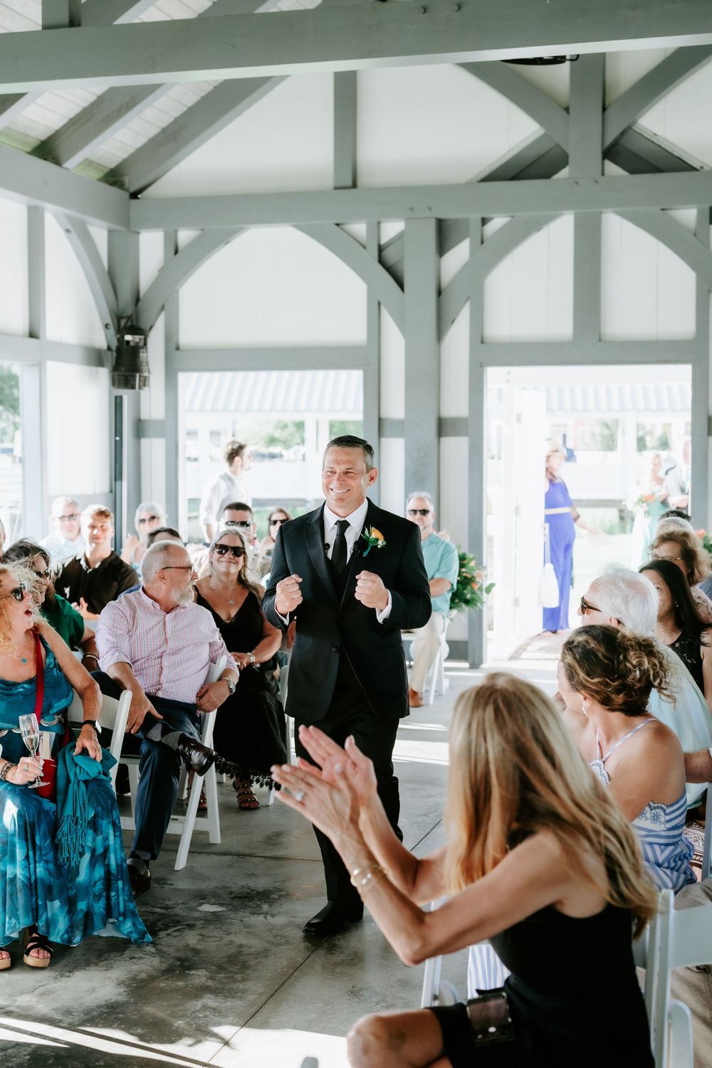 Groom in black suit walks down aisle at outdoor wedding ceremony as guests applaud and smile.