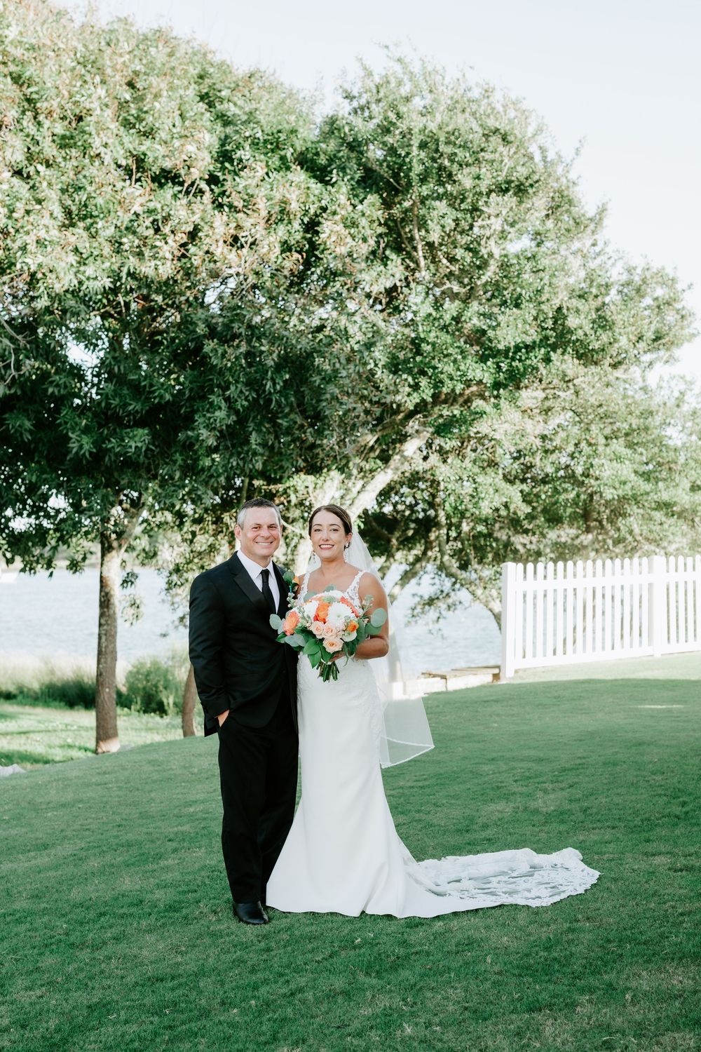 Bride and groom pose together on green lawn under large tree near white fence and waterfront on wedding day.