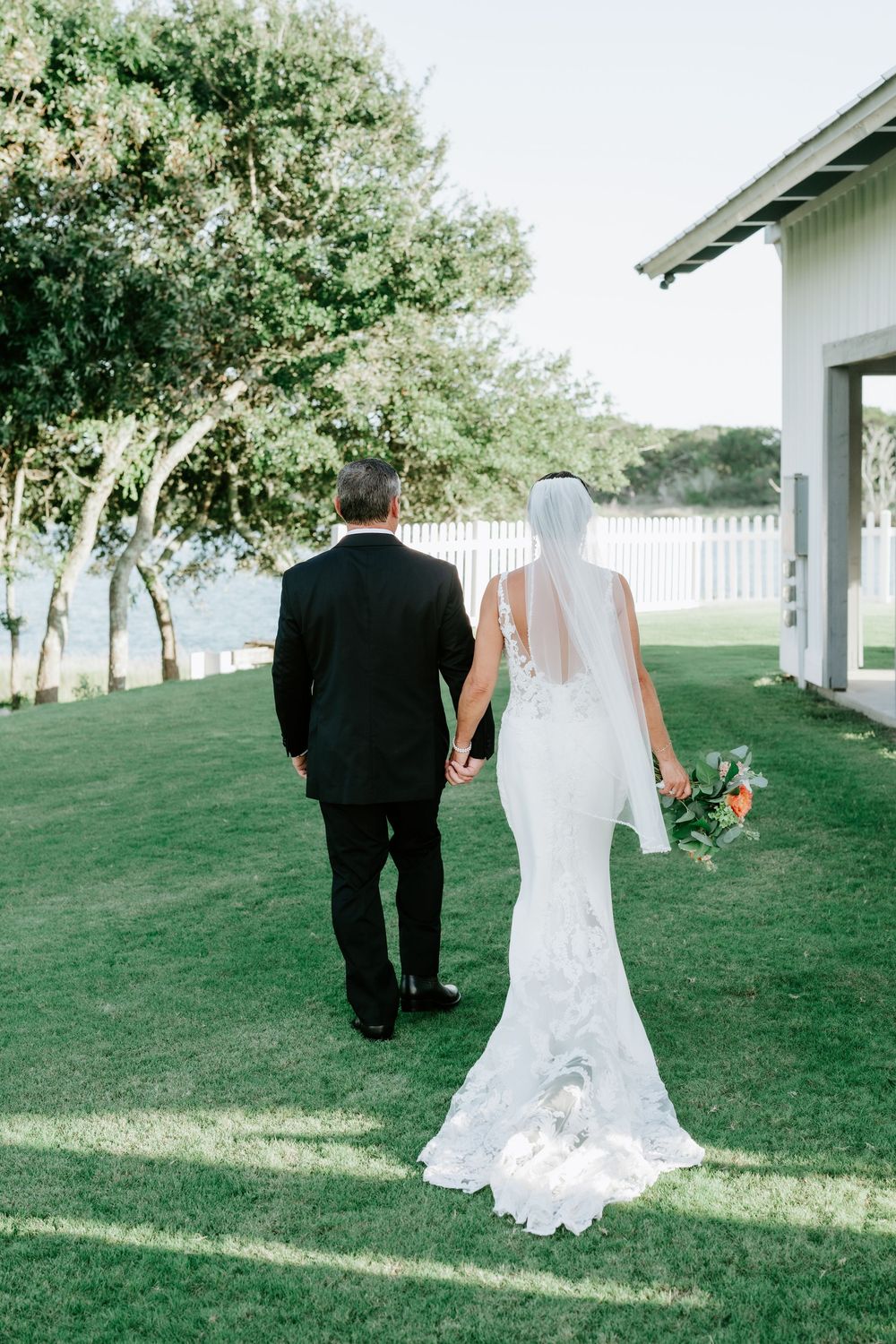 Bride in white lace gown and veil walks with groom in black suit across green lawn toward waterfront.