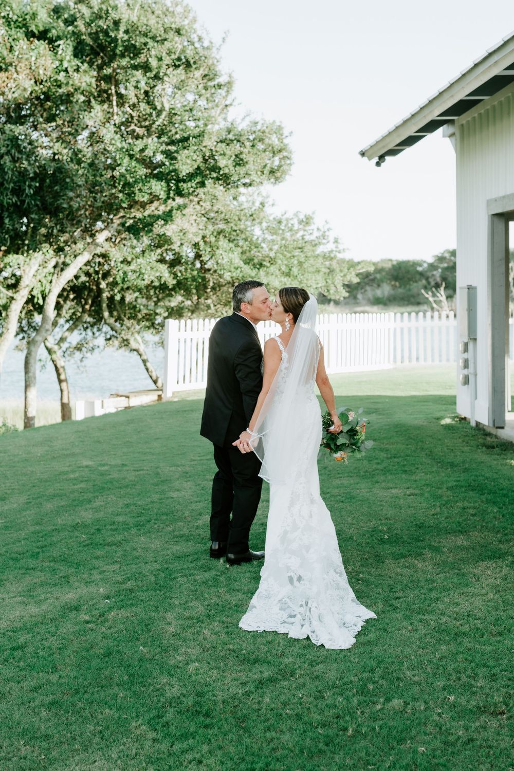 Bride and groom share a kiss on green lawn near white fence and waterfront, bride holding colorful bouquet.