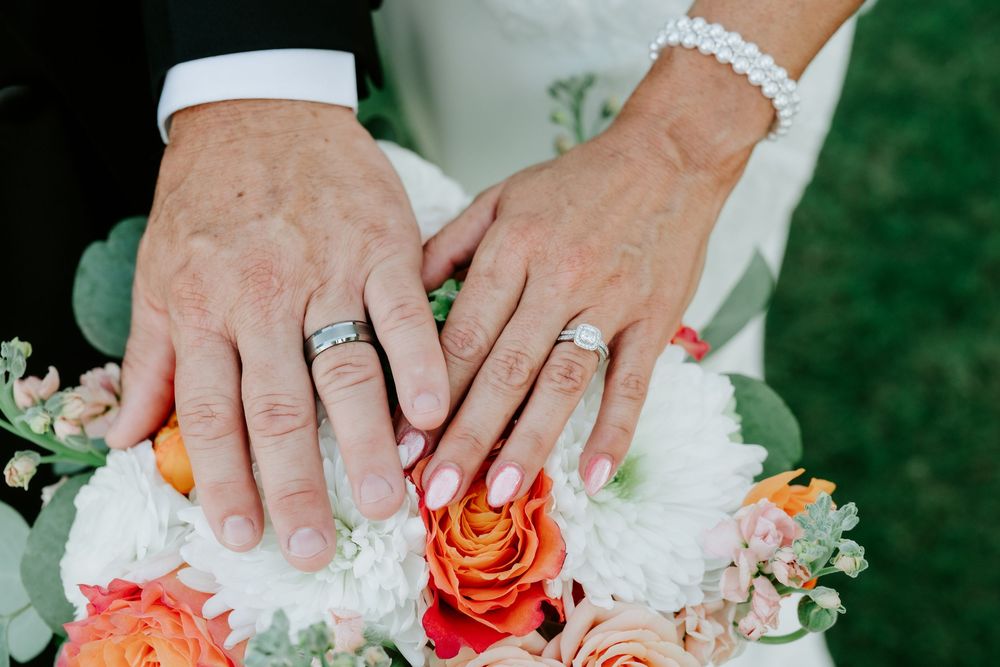 Newlywed couple displaying wedding rings on hands resting over a colorful bridal bouquet with roses.