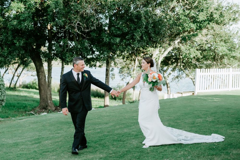 Bride and groom hold hands walking on green lawn under trees in outdoor wedding portrait.