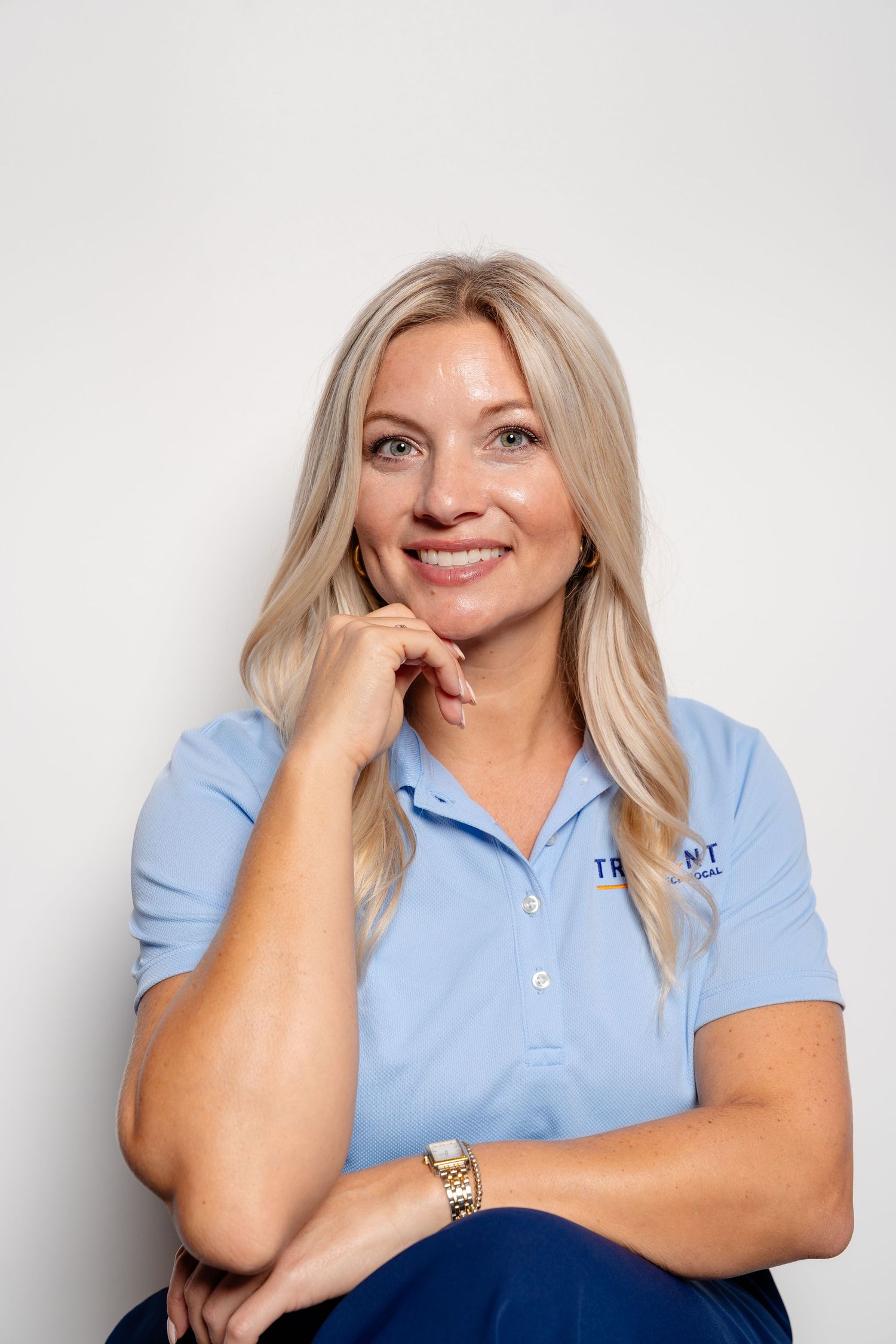 Smiling blonde woman in light blue Trident polo shirt poses confidently against white background.