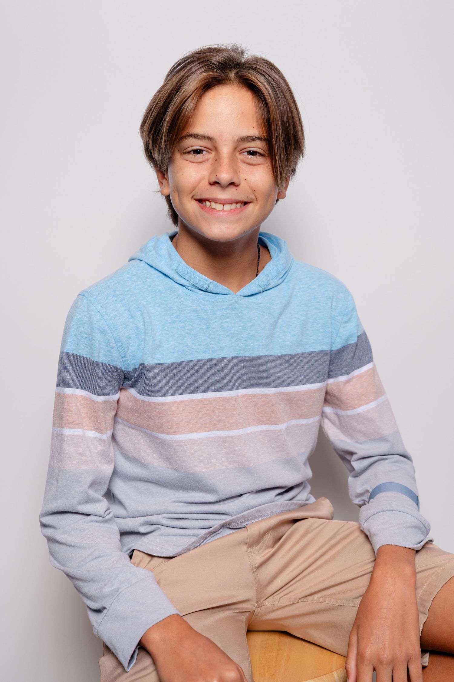 Smiling preteen boy with brown hair wearing a striped blue hoodie and khaki shorts, sitting against a white background.