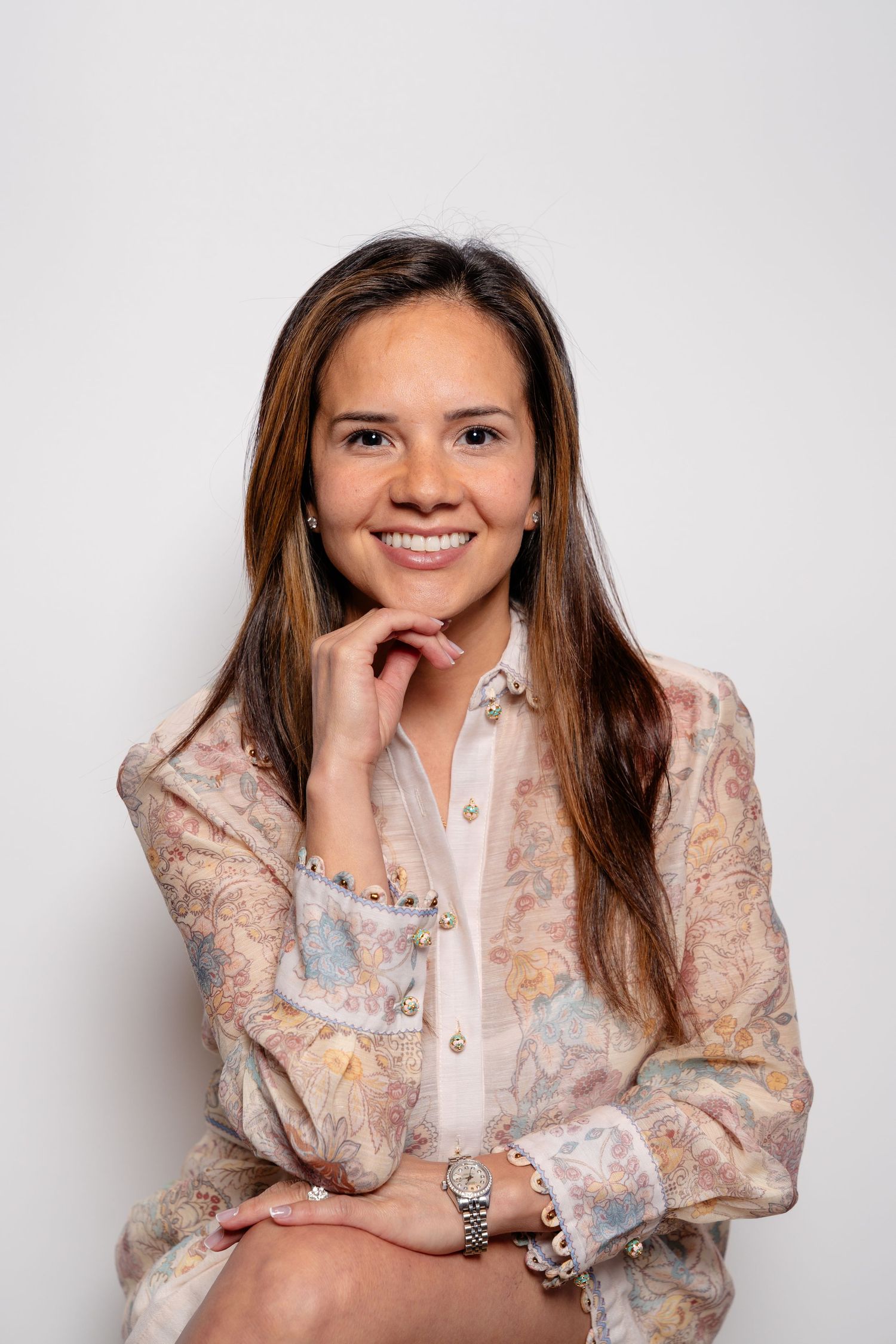 Smiling young woman in floral blouse with hand on chin, posing confidently against white background.