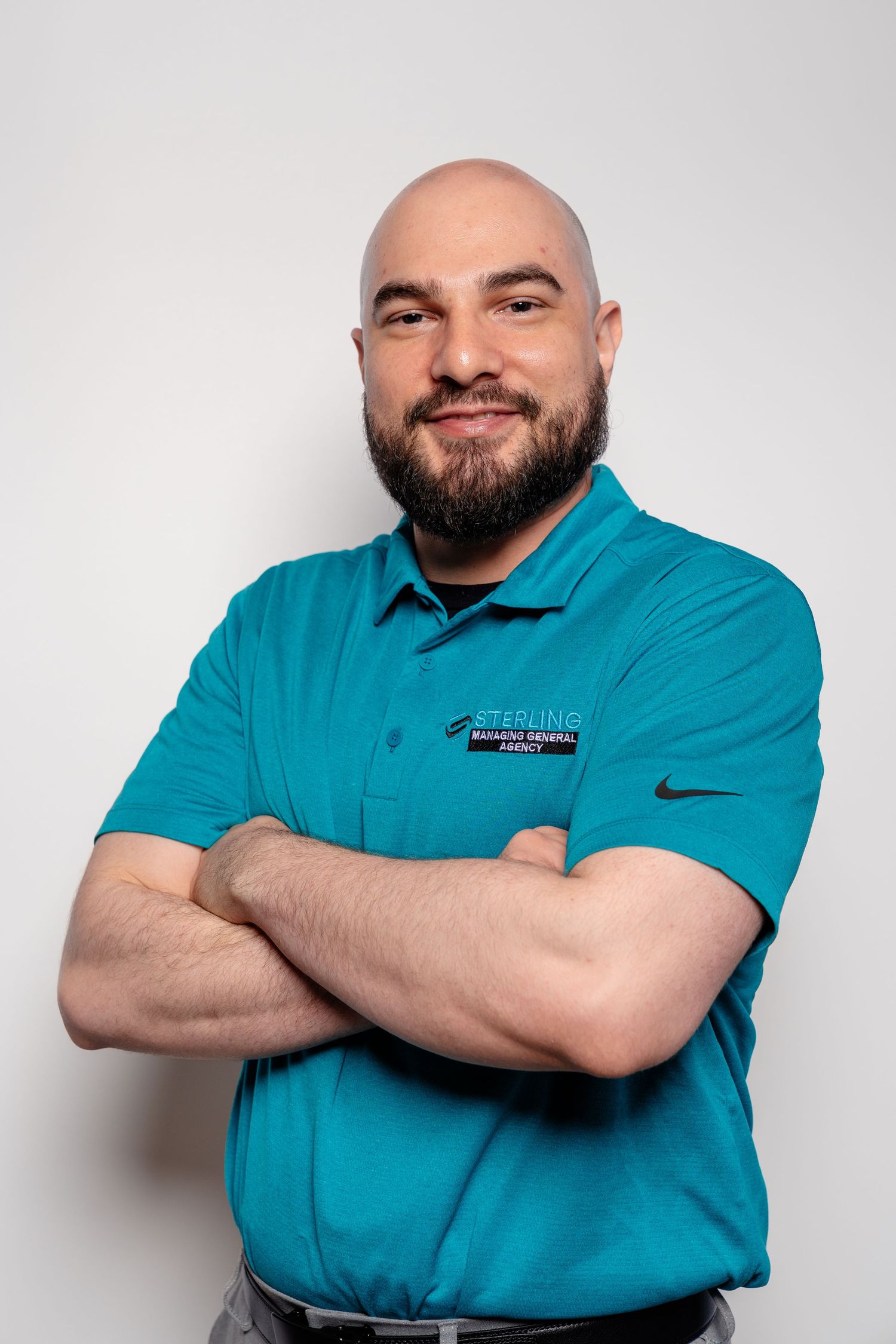 Bald bearded man in teal Nike polo shirt stands with arms crossed smiling against white background.