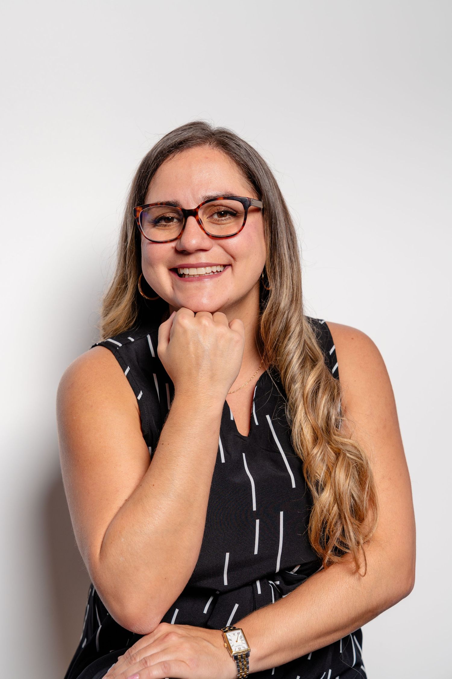 Smiling woman with glasses and long hair wearing a black patterned sleeveless top, posing with hand on chin.