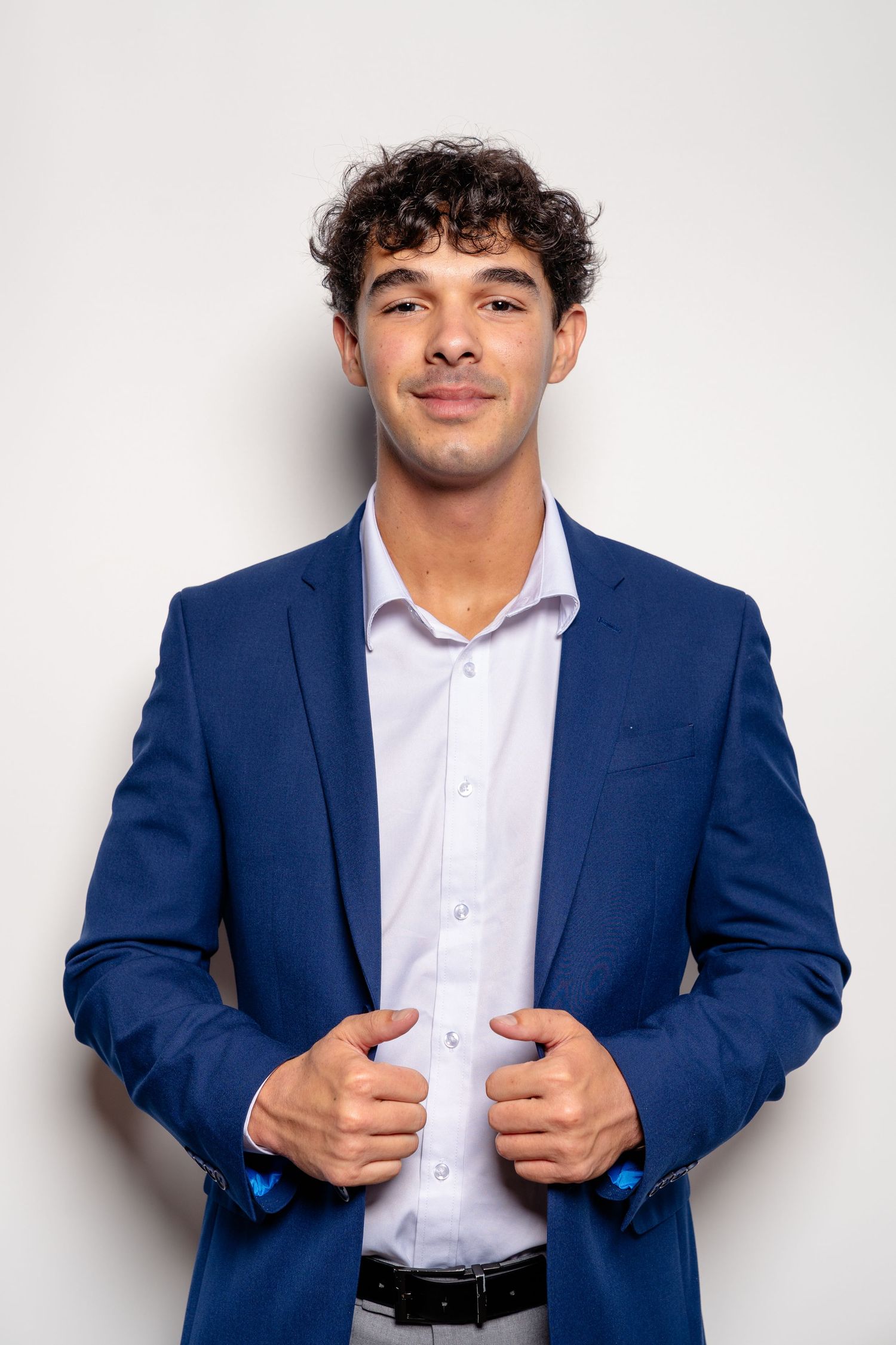 Young man in blue blazer and white shirt smiling confidently against a light gray background.
