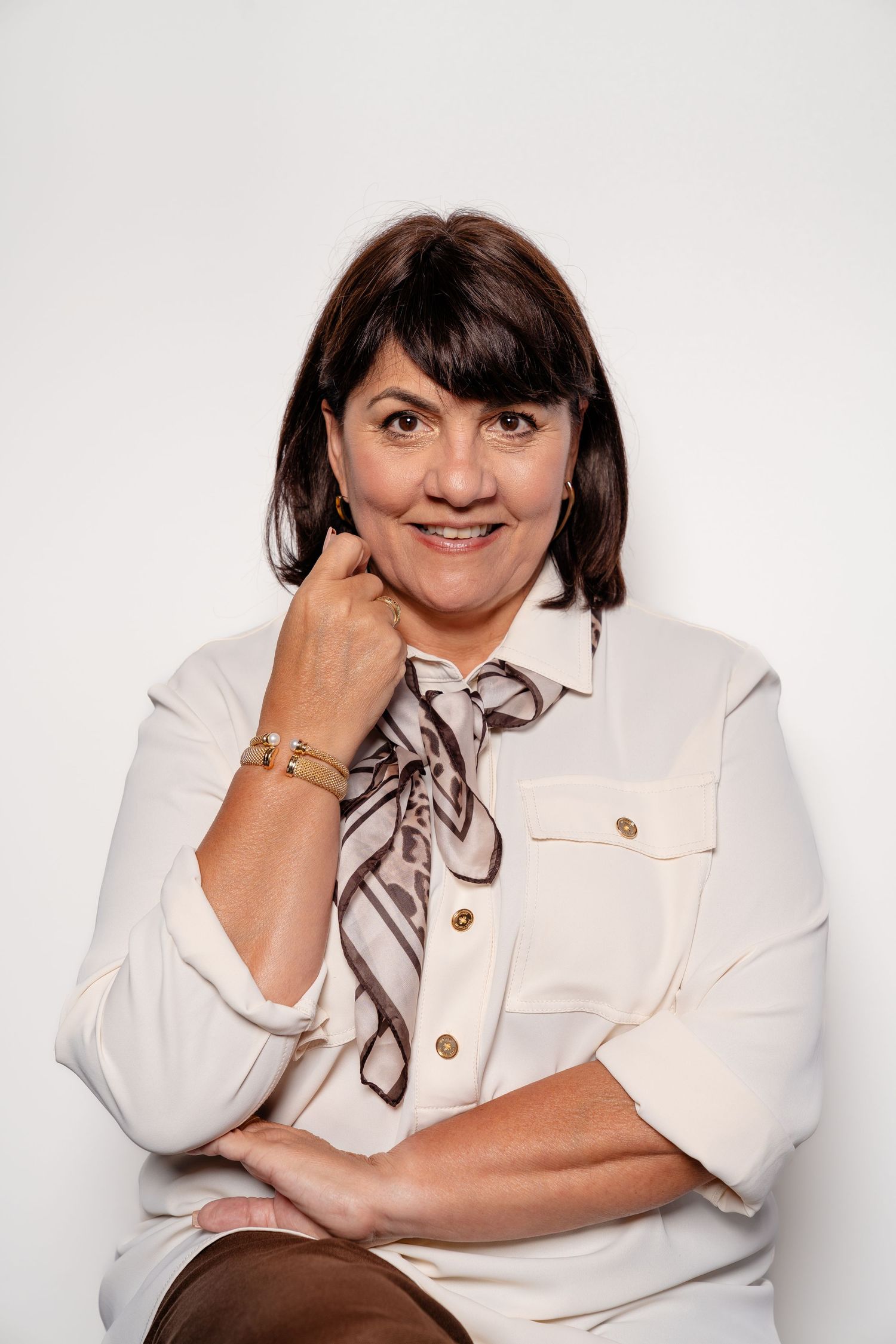 Smiling middle-aged woman with dark hair wearing a white blouse and silk scarf, posing against a white background.