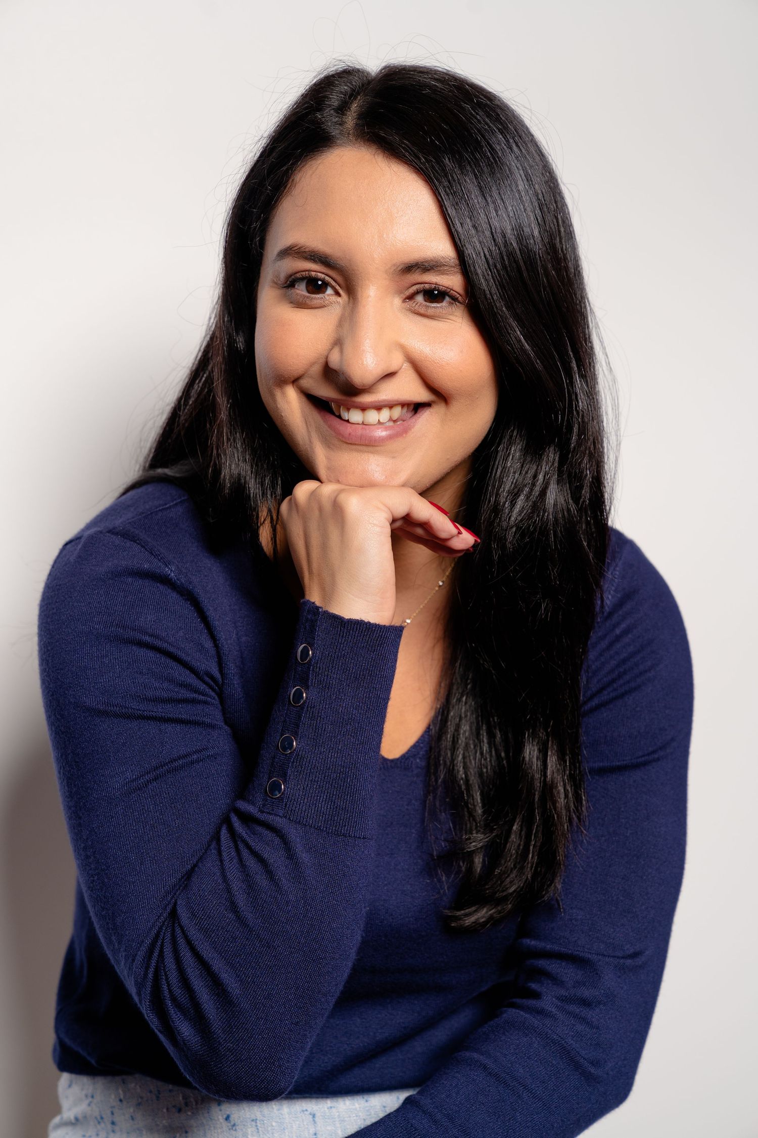 Smiling woman with long dark hair wearing a navy blue top, posing with hand under chin against white background.